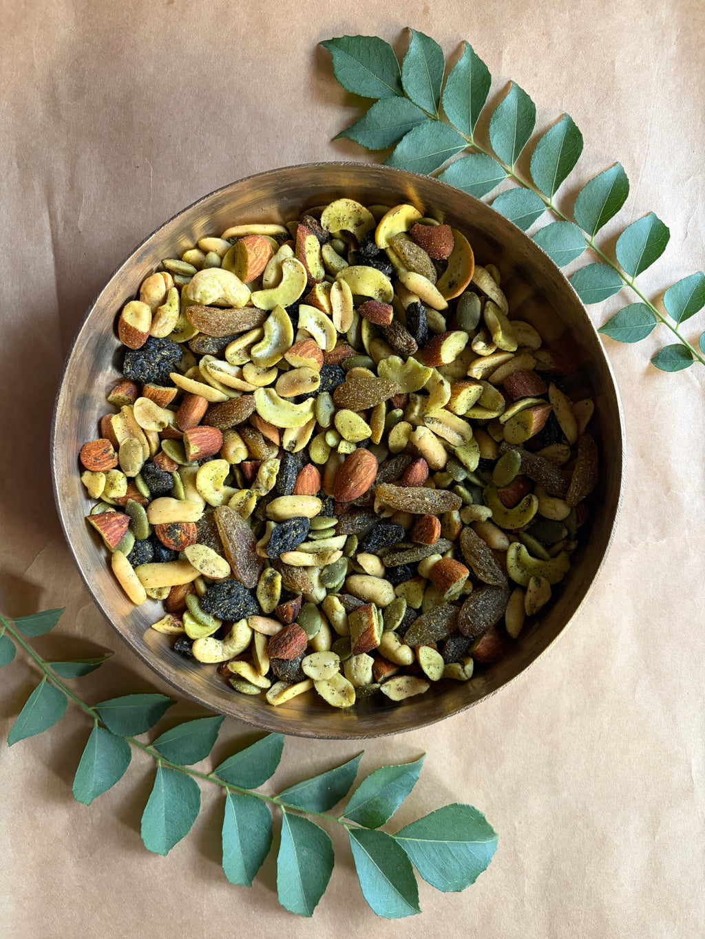 Assorted nuts and seeds trail mix in a bowl on a wooden surface with curry leaves.